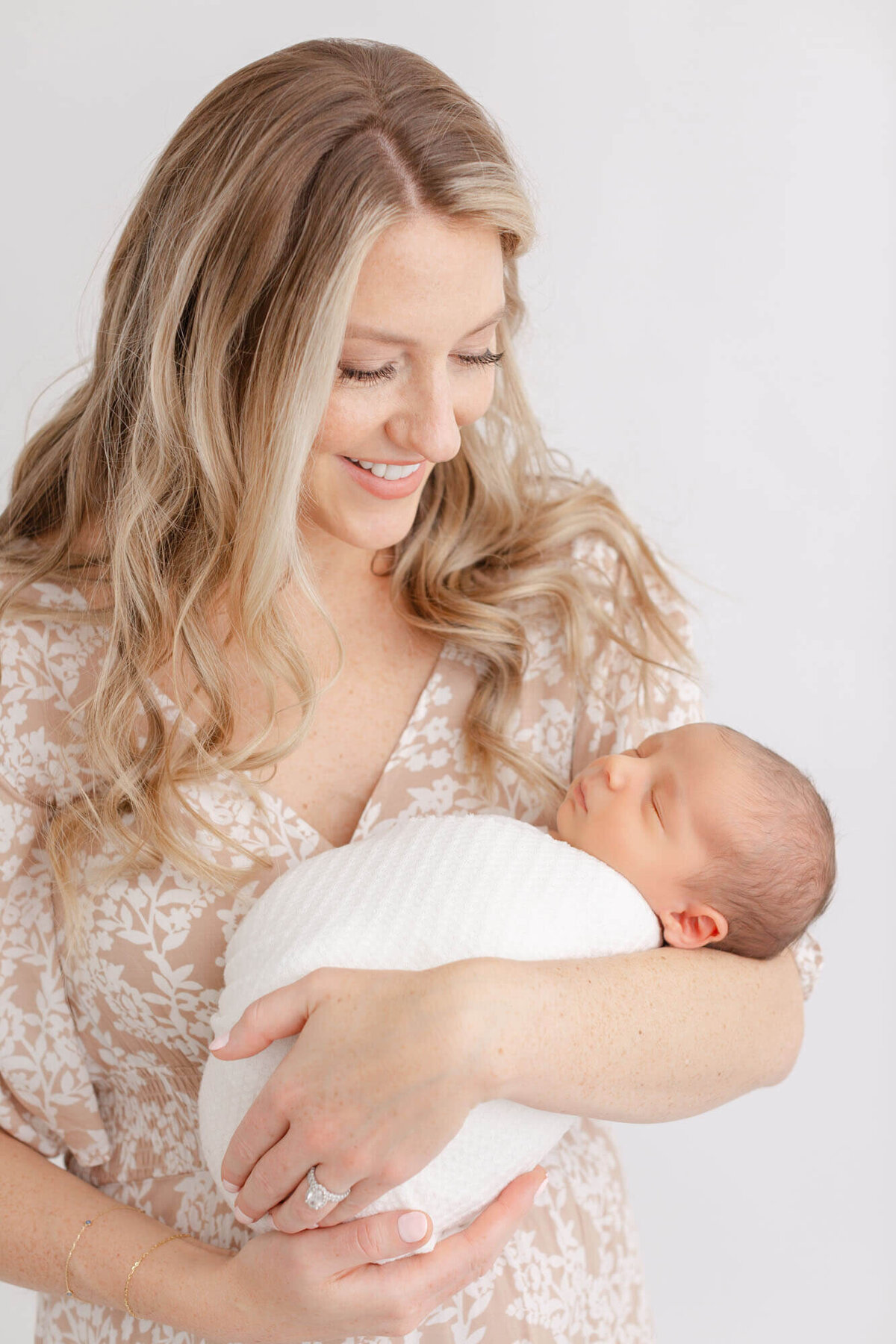 Mom in beige floral dress holding her baby in her arms and smiling down at him.