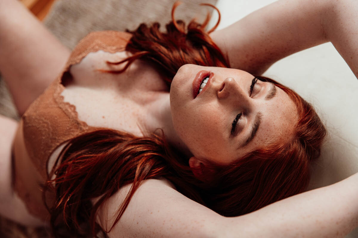Boudoir portrait of redhead leaning against couch with eyes shut.