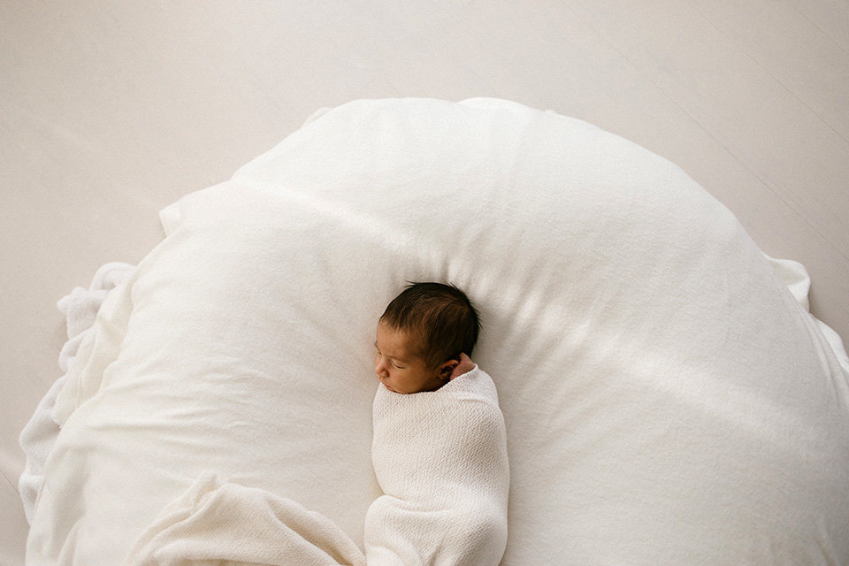 Newborn baby is laying wrapped on a white bean bag chair with sunlight streaming in