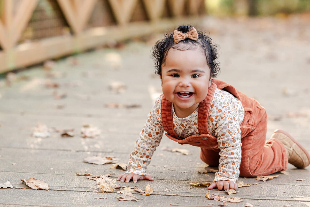 A toddler wearing orange overalls and a matching bow in her hair crawls along a bridge in  a park in Virginia Beach.