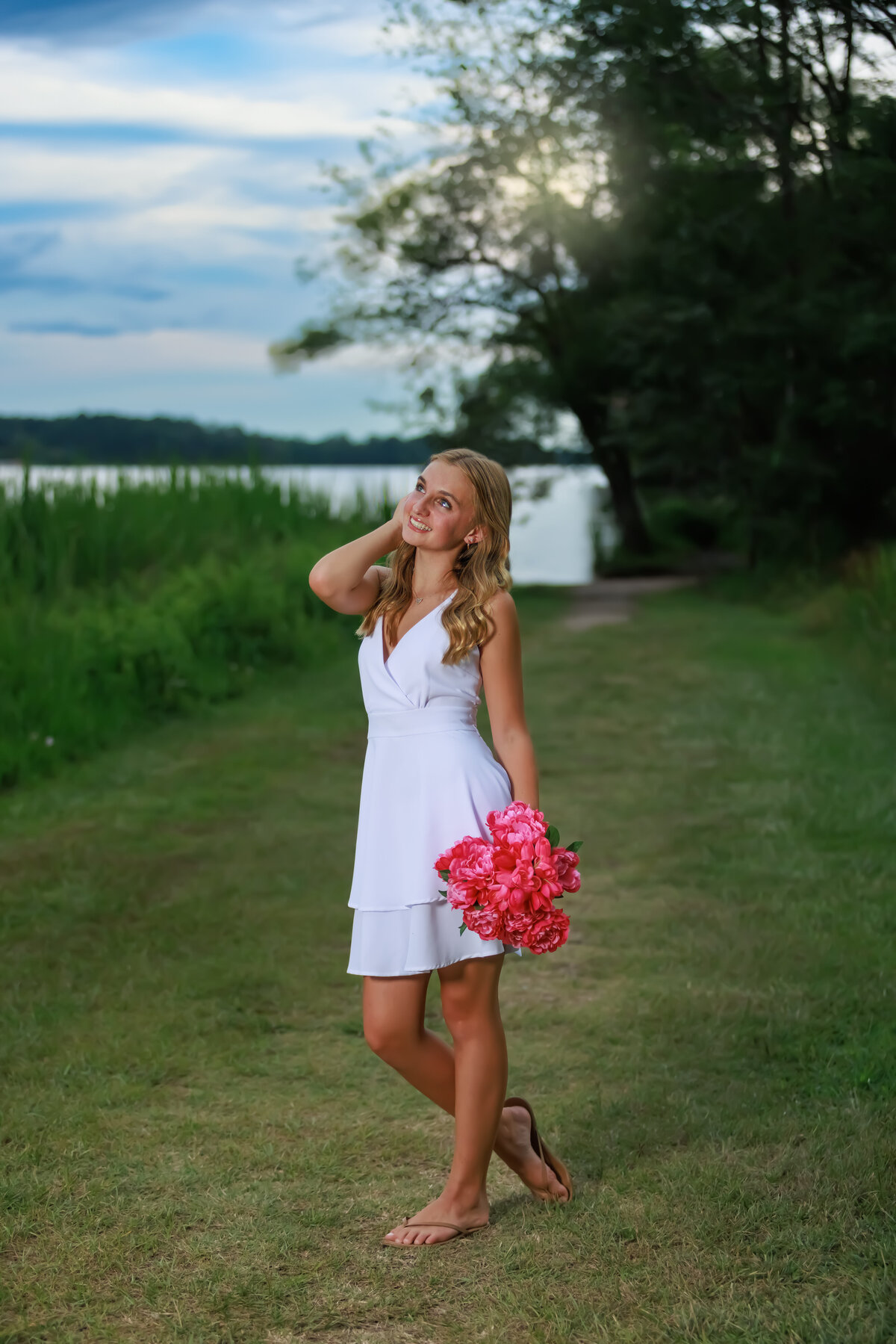 Raleigh Teen by a lake at sunset wearing a white dress holding pink flowers.
