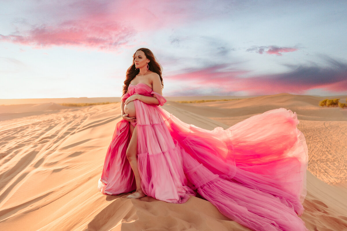 mother wearing bright pink tulle maternity gown holding her baby bump in the desert