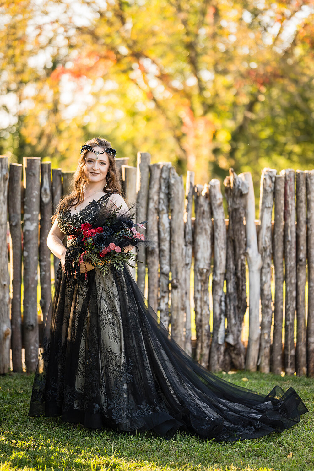A lesbian bride wearing a black wedding dress and holding a DIY black and red wedding bouquet stands and smiles in the backyard of an Airbnb at golden hour in Roanoke, Virginia for a formal portrait.