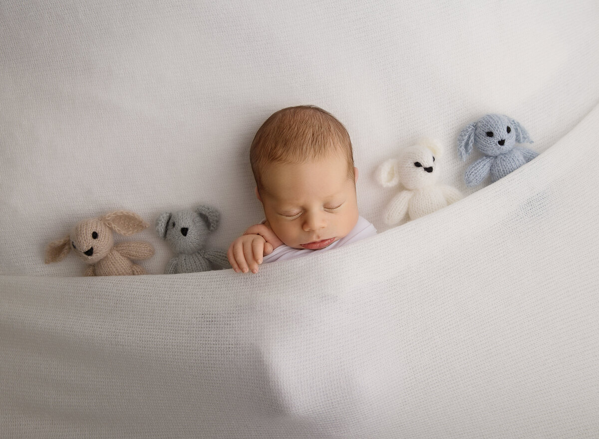 Baby boy is laying underneath white stretch fabric with his hands peeking out. Beside him there are a series of mini felt teddy bears. Captured by top Brooklyn newborn photographer Chaya Bornstein