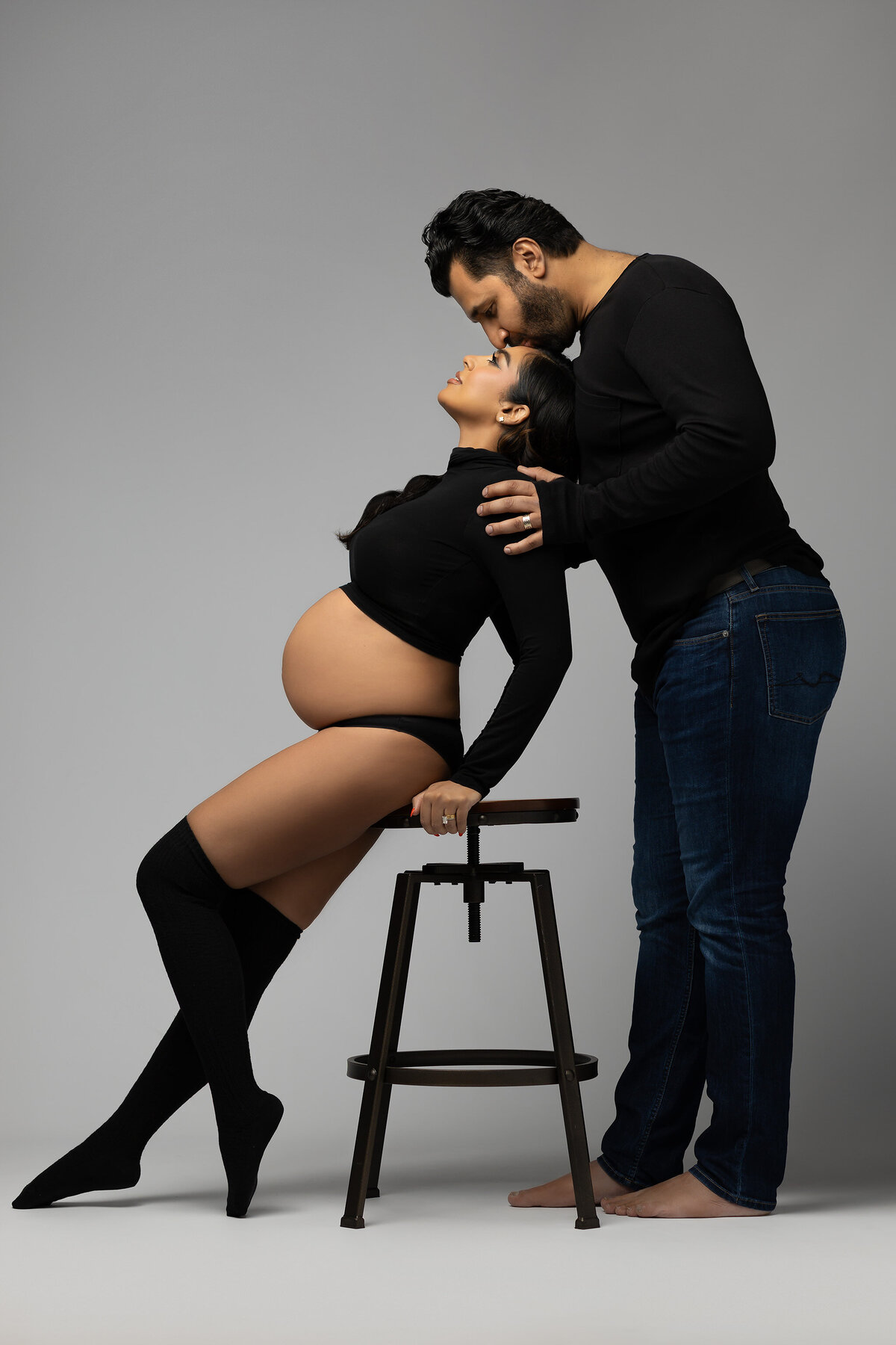 A woman in black clothing leans on a stool while a man gently kisses her forehead. The scene conveys love and tenderness in a studio setting. Captured by DFW Maternity Photographer Lindsay Walden.