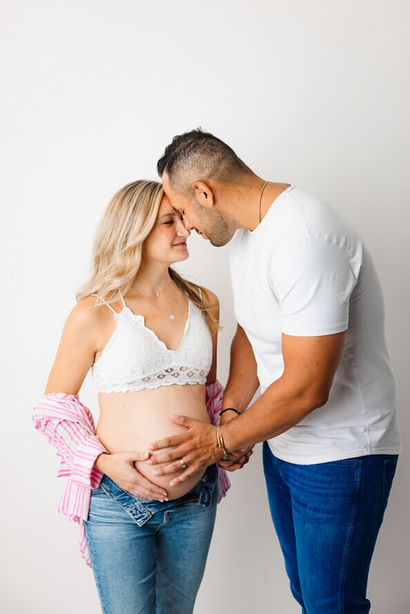 A husband and pregnant wife embrace on a white backdrop.