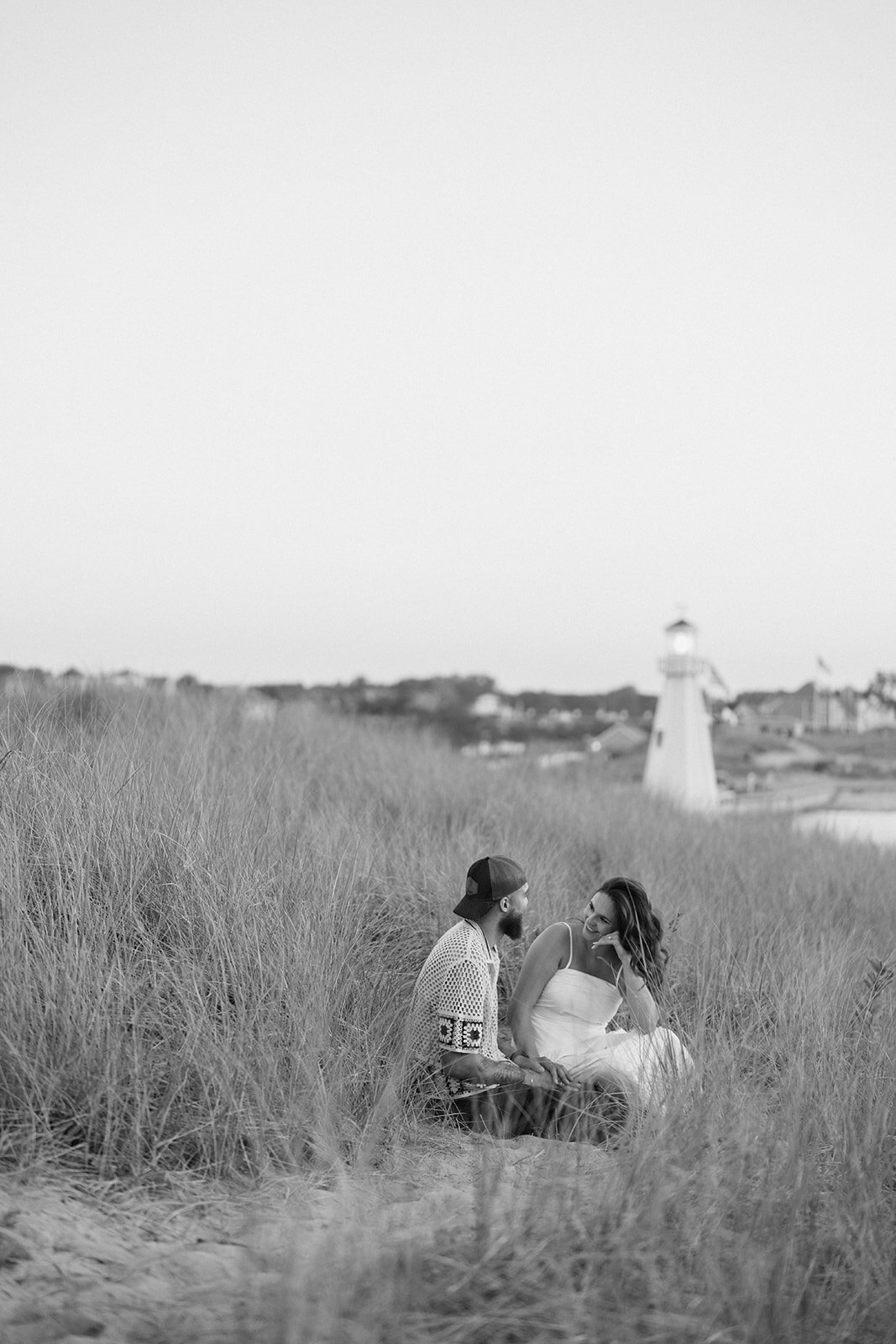 Black and white engagement photo of couple in the dunes with New Buffalo lighthouse behind them