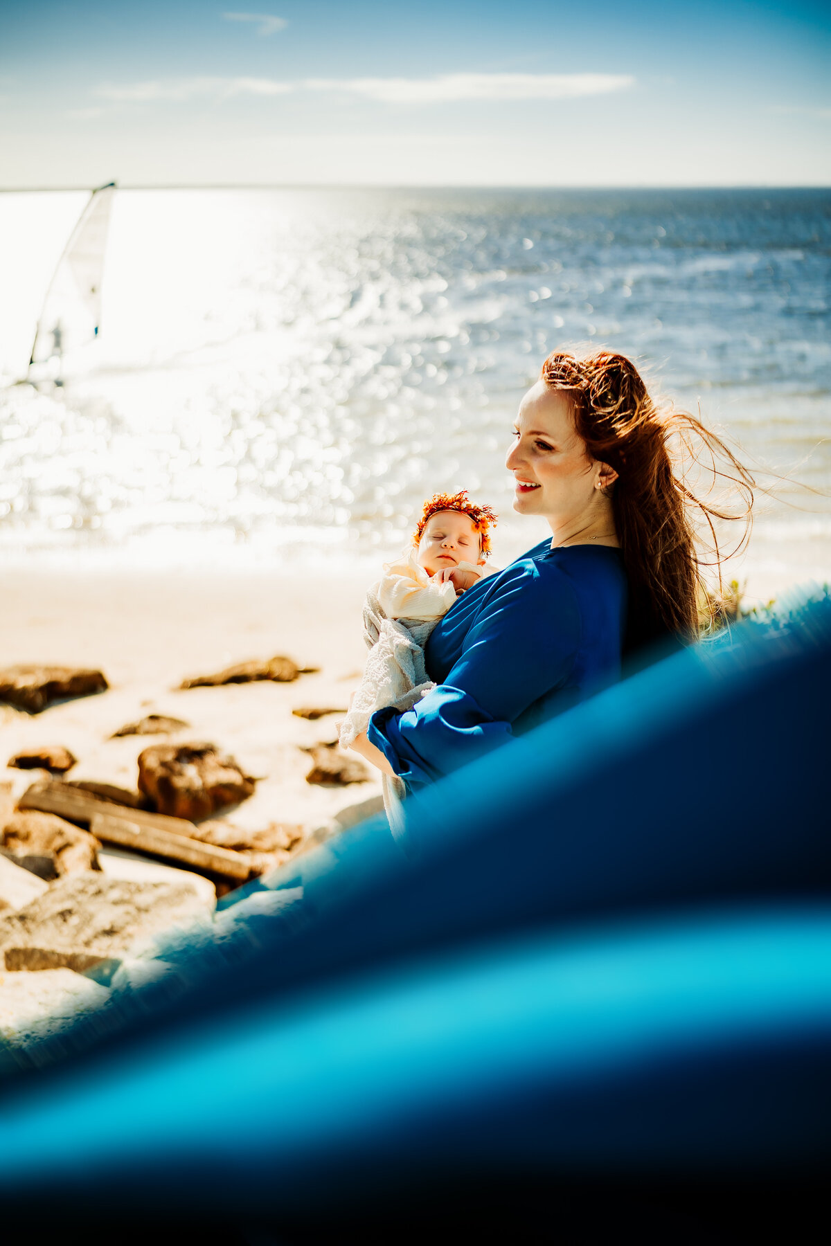 a mom in a stunning flowy blue dress holding her newborn baby at the beach at sunrise