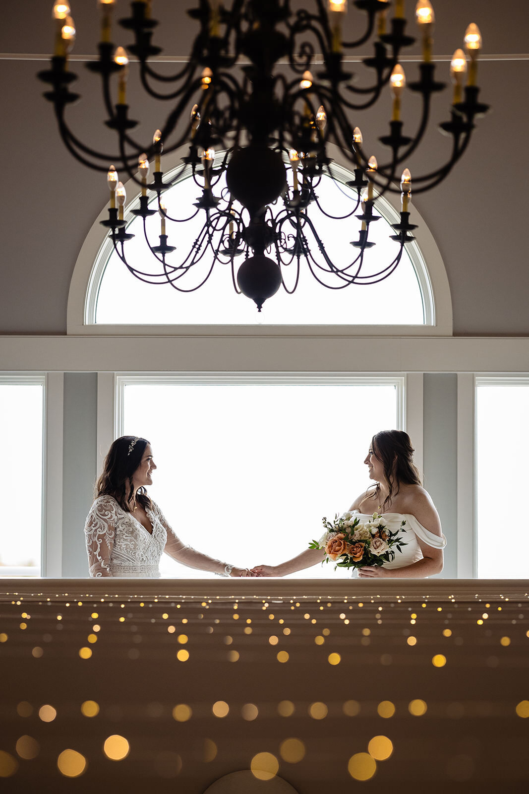 Two brides facing each other holding hands, with a chandelier above and twinkling lights below, creating a romantic atmosphere.