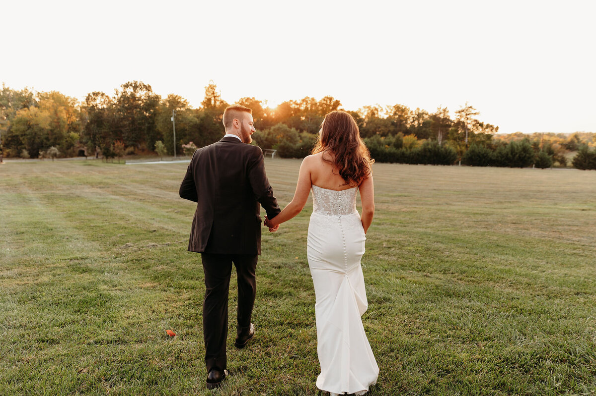 A couple in formal attire strolls hand in hand across a grassy field at sunset, perhaps reflecting on their perfect day with one of the exquisite wedding packages Virginia offers. The woman wears a white dress, and the man is in a dark suit. Trees line the horizon as the sun casts a warm glow over their serene moment.