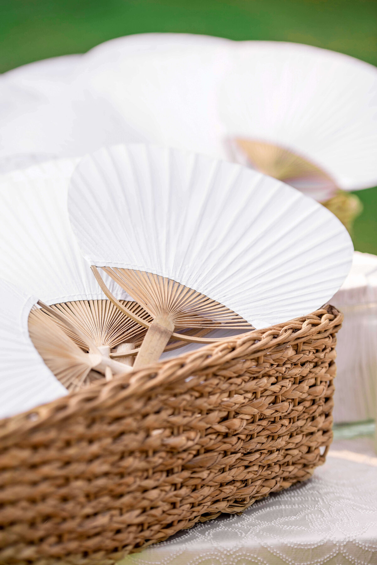 A woven basket filled with white paper fans, each with a wooden handle, sits on a lace-covered table. The fans are partially open and the setting appears tranquil, possibly outdoors.