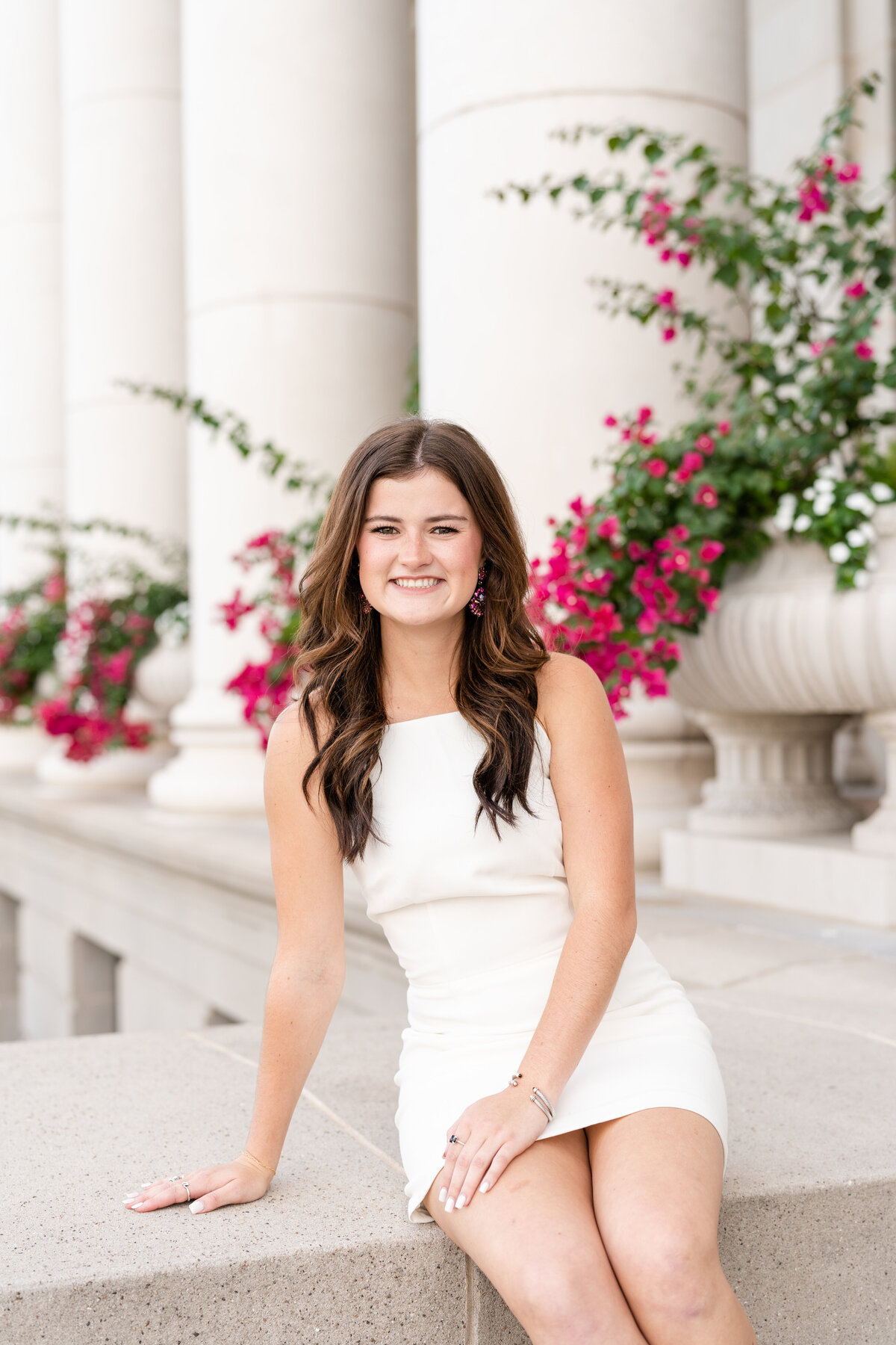 Texas A&M senior girl sitting in front of Administration Building while wearing white dress and smiling with pink flowers behind her