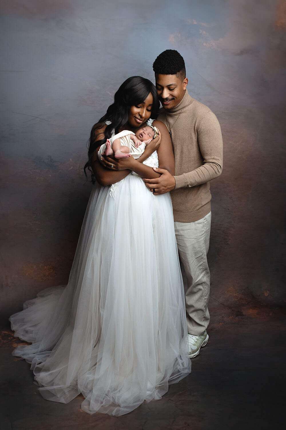 Happy new parents stand in a studio cradling their sleeping newborn baby taken by a New Orleans Newborn Photographer