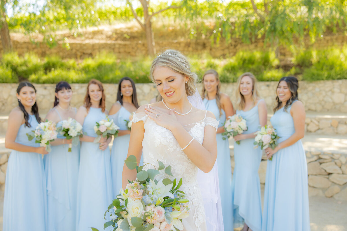 Bridesmaids in blue dresses stand in background looking at bride in the front who smiles and looks down at her ring and bouquet of flowers. Photo taken by wedding photographer from Sacramento, philippe studio pro.