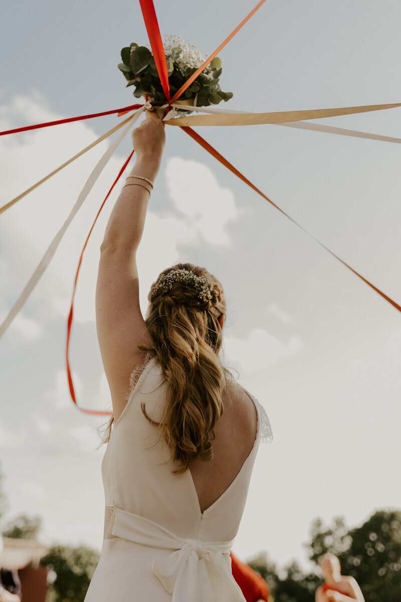 Mariée de dos tenant à bout de bras son bouquet et des rubans de différentes couleurs. Photo de mariage réalisée en Vendée.