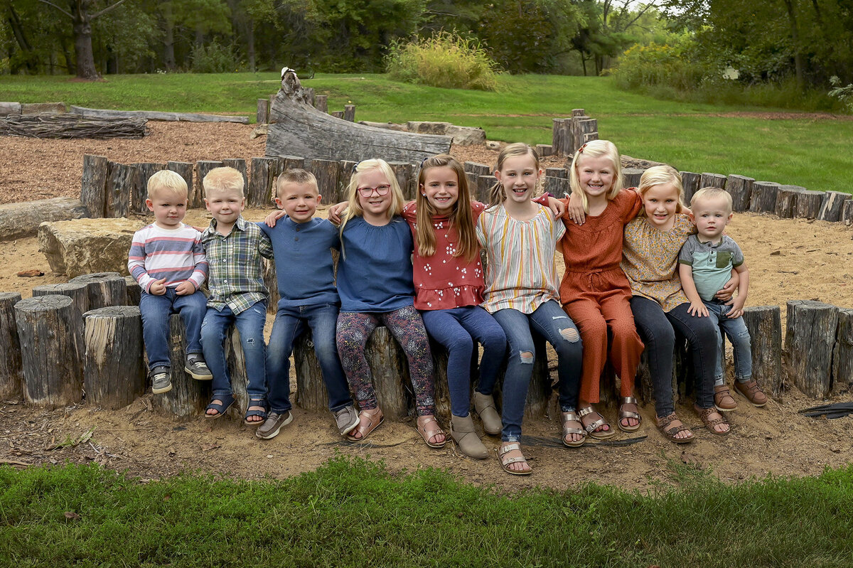 9 children sitting together on a large wooden log in an outdoor setting with trees and greenery in the background