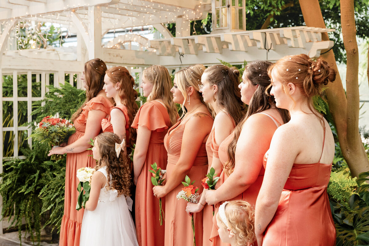 A lineup of bridesmaids and a flower girl stand elegantly at the altar, dressed in burnt orange gowns, holding flowers, and adorned with delicate pearl hairpins.