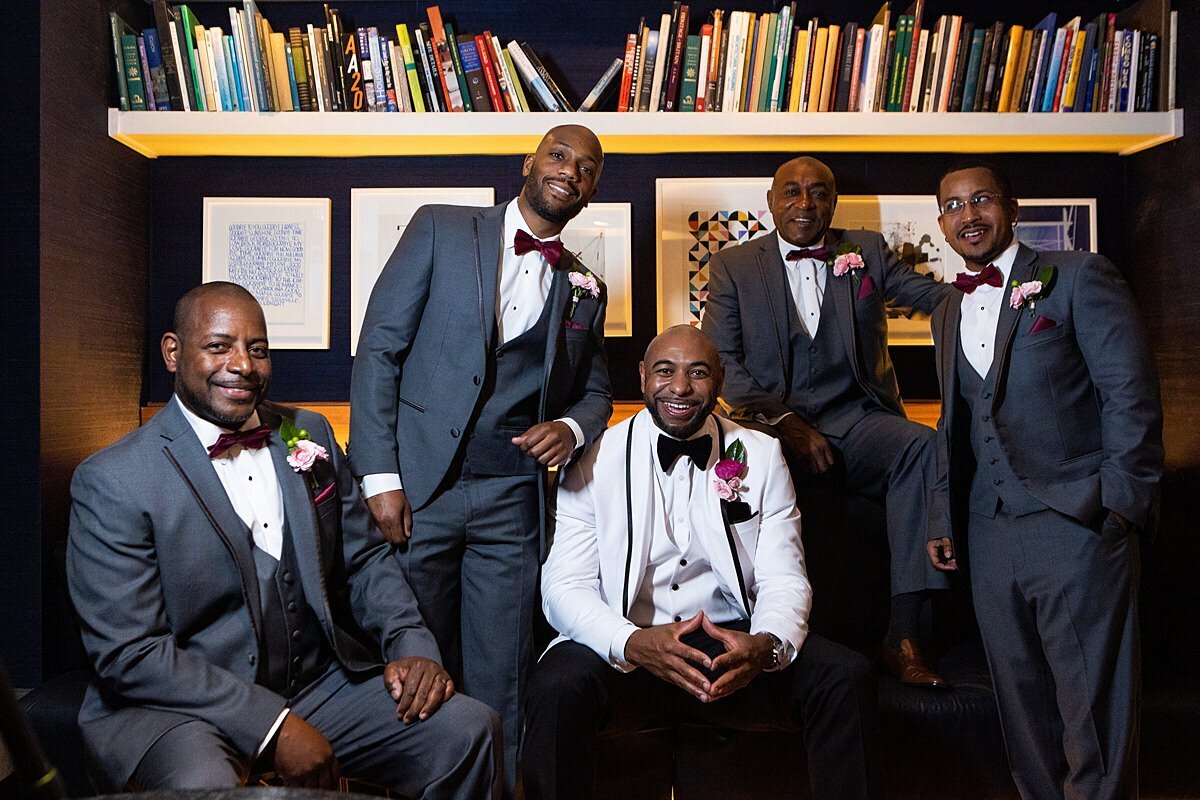 Groom wearing a white tuxedo and groomsmen wearing dark grey tuxedos sitting in a library