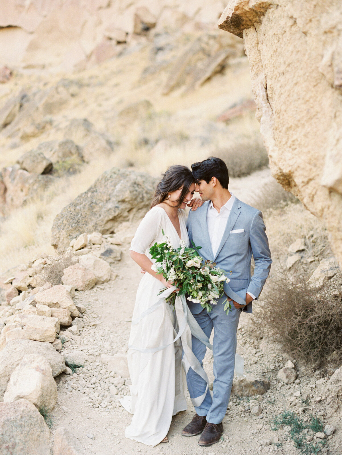 Bridegroom in light blue suit whispers to his newly married wife at a rocky backdrop.
