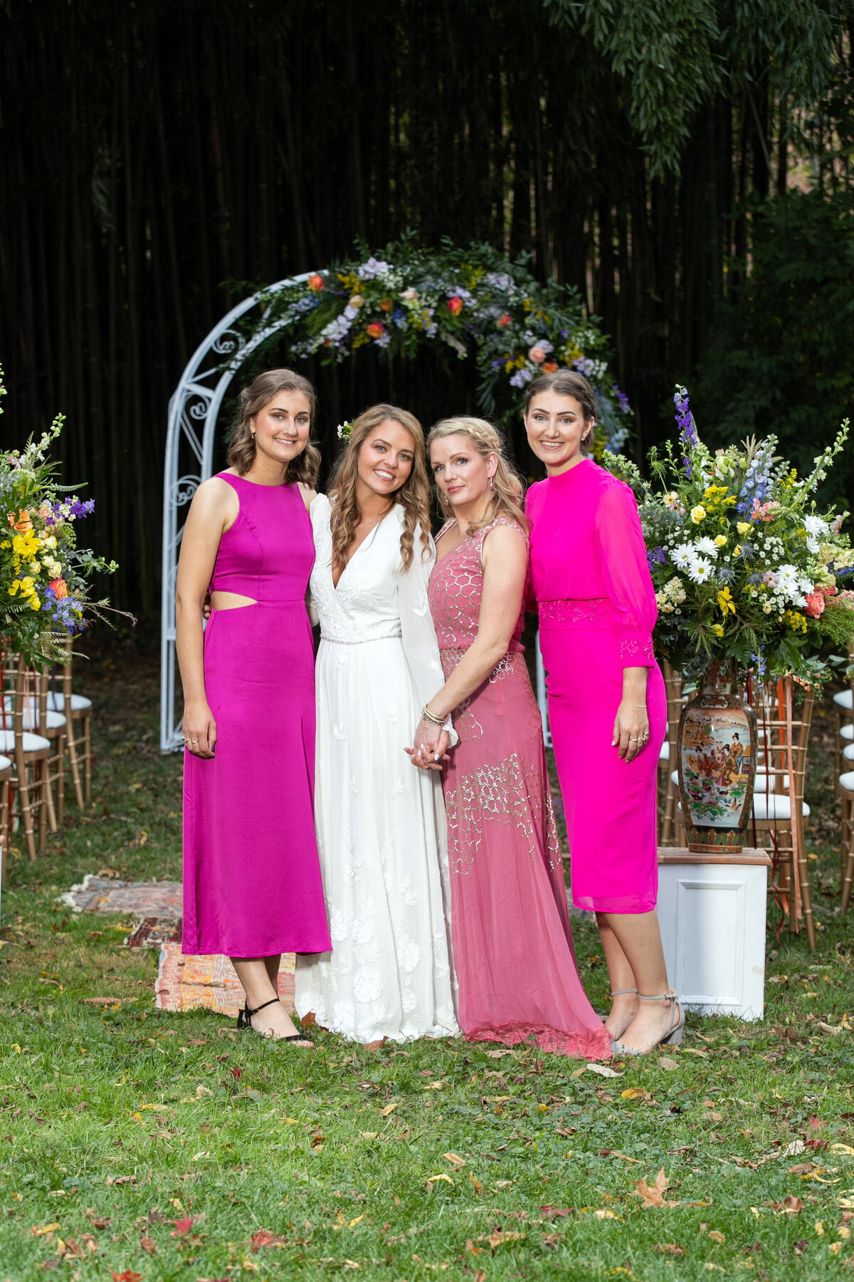 Three bridesmaids wearing pink stand under a floral arch with the bride