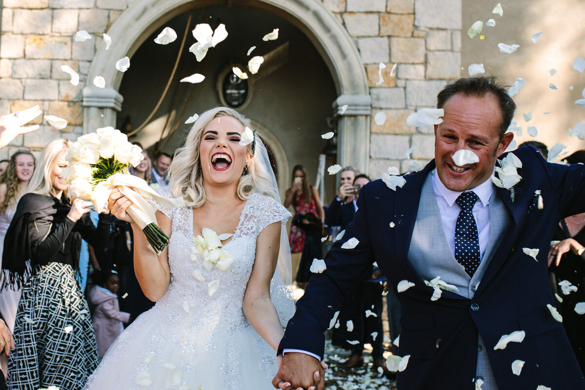 Newlyweds are laughing spontaneously while being showered with white rose petals. 