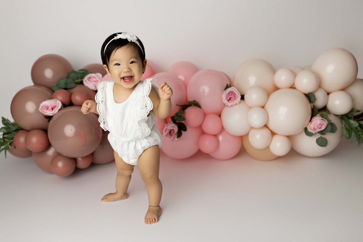 Baby girl standing in a white lace romper and matching headband smiling at the camera. A pink and dusty rose balloon garland is behind her. Photo taken by top London, Ontario baby and cake smash photographer Amy Perrin-Ogg