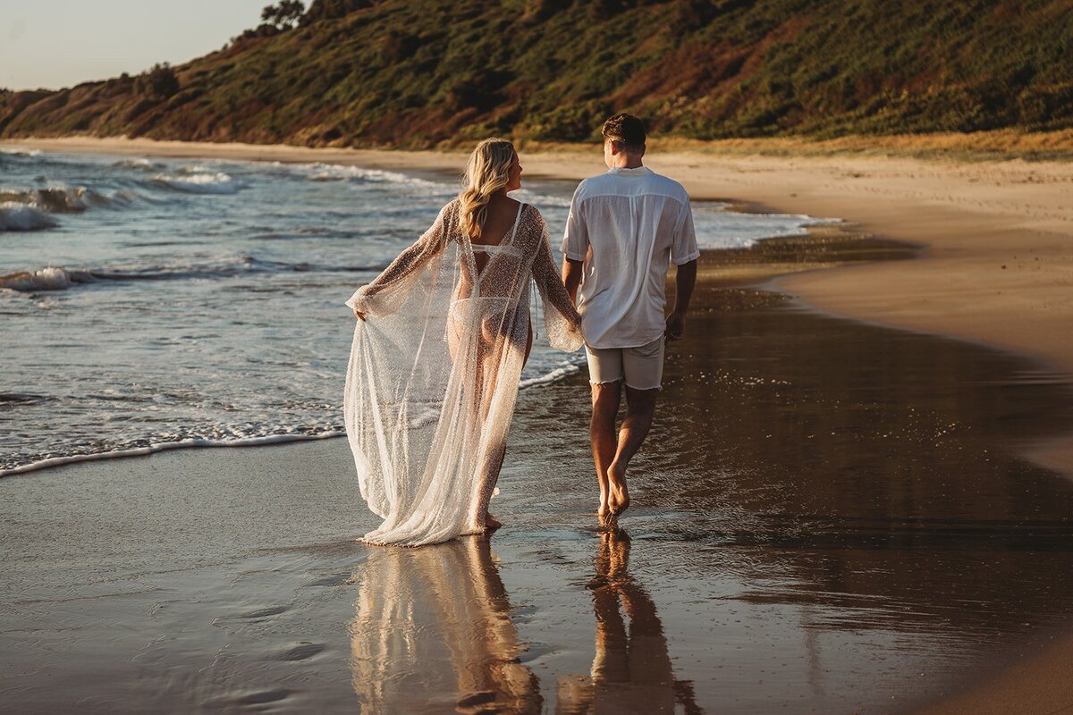 A couple walks hand-in-hand along the shoreline of a sandy beach at sunset, with the woman wearing a sheer cover-up and the man dressed in a white shirt and shorts. Capturing moments like these is what makes an NSW family photographer's job truly special.