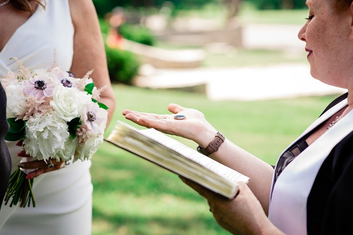 A detail image of the wedding officiant offering the wedding bands to the bride. In one hand she is holding a book and in the other hand, the wedding bands. The officiant is wearing a black dress with a white officiant sash as she looks down at the rings in her hand. The bride is off to the left wearing a white fitted dress with a v-neckline holding a bouquet of white and blush flowers.