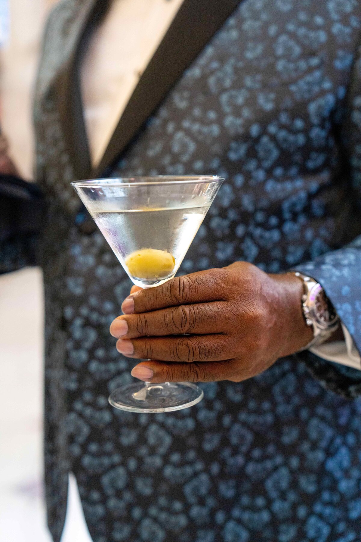 A person holds a martini glass with a green olive. They wear a patterned dark blue suit with a black lapel and a shiny silver watch.