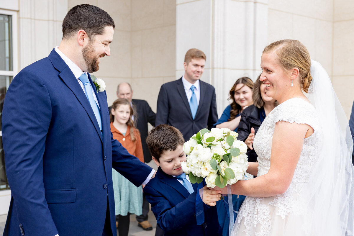 Bride and groom smile warmly while a young boy touches the bride’s bouquet, surrounded by family members in front of the temple.