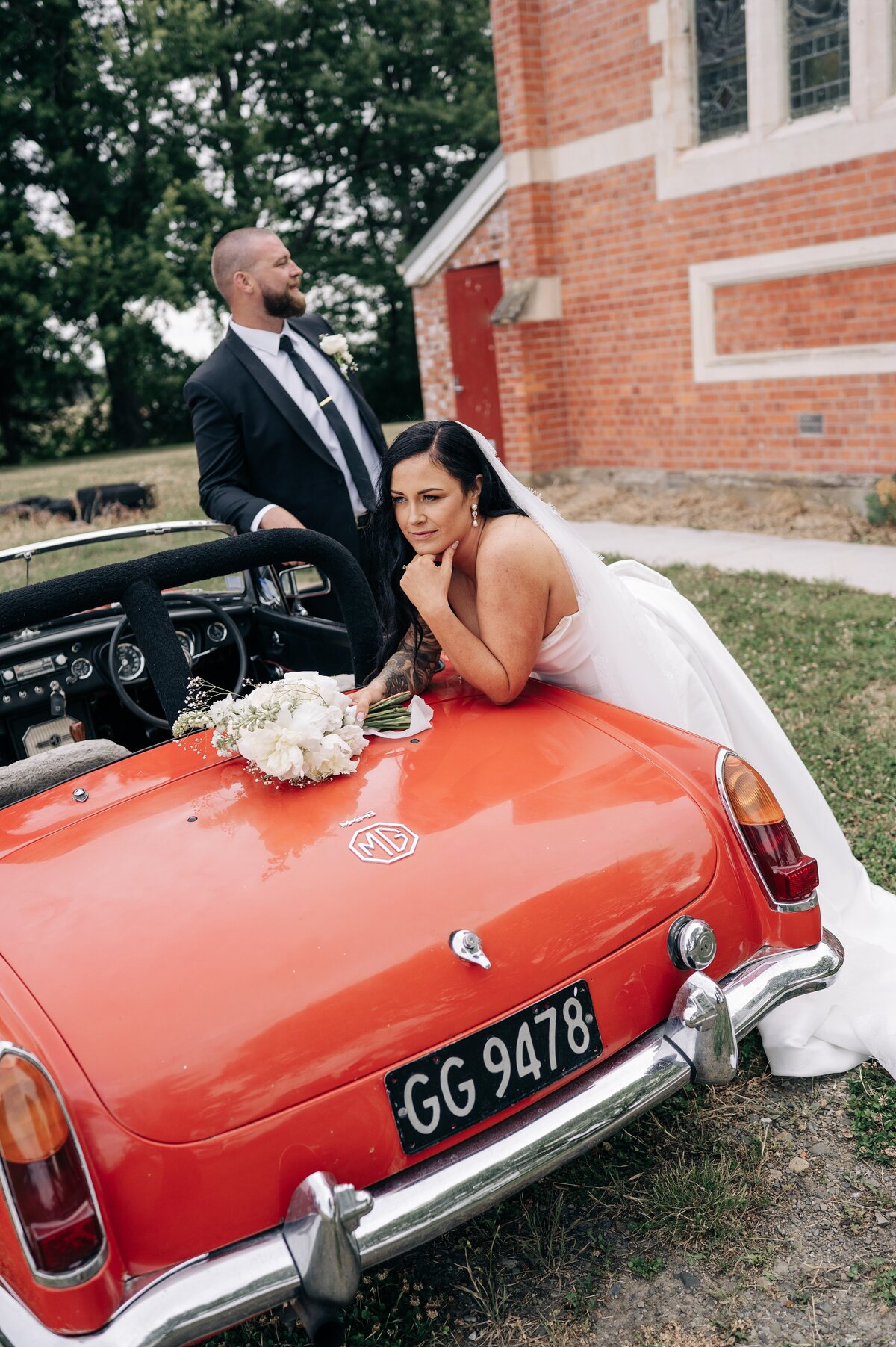 bride leans on the back of a red MG convertible by a church at their christchurch wedding