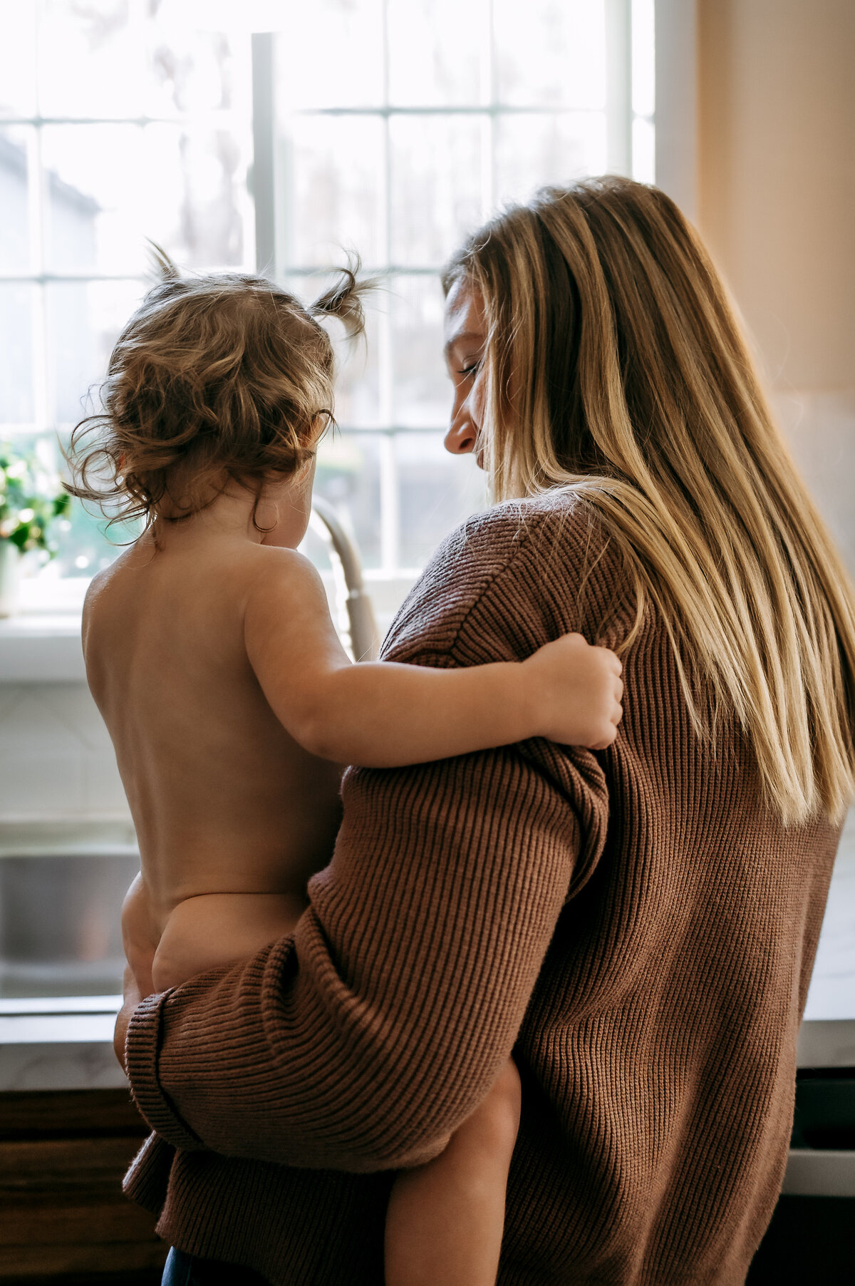 Mom holding her toddler by the window during a soft, natural in-home motherhood session.