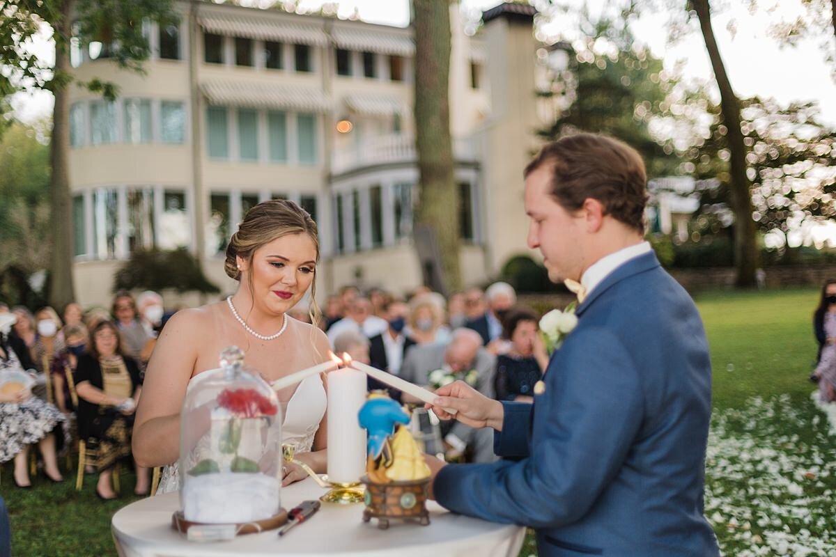A bride in a strapless wedding gown with a pearl necklace and plunging neckline hold a white taper candle over a tall white pillar candle as the groom, also holding a white taper candle , wearing a blue suit with a white shirt lights the unity candle. A Beauty and the Beast music box and an Enchanted Rose sits on the table next to the candles. The Estate at Cherokee Dock is in the background.