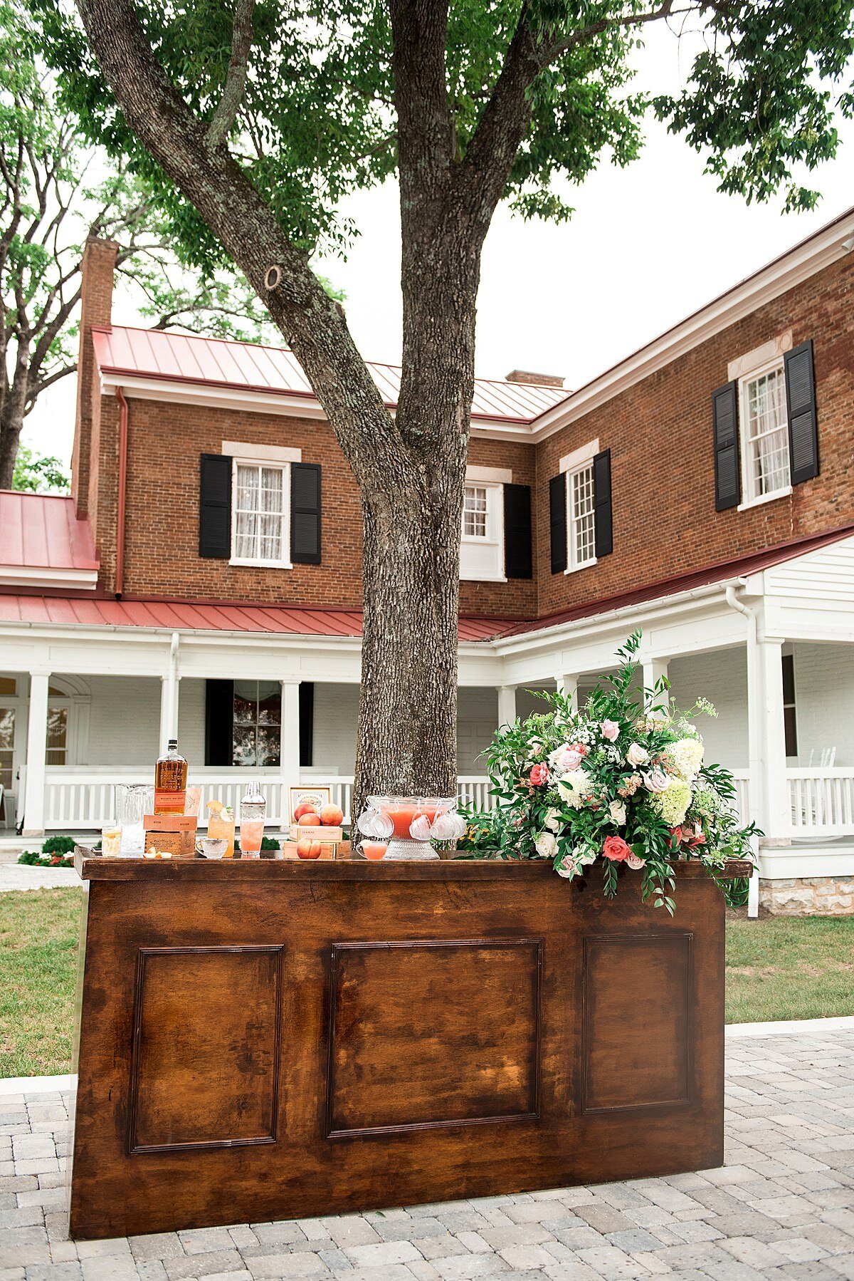 The dark wood bar in the courtyard of Ravenswood Mansion is decorated with a large centerpiece of greenery, pink and peach , white and ivory flowers as well as a selection of signature summer mocktails and signature summer cocktails as well as flavored wedding teas.