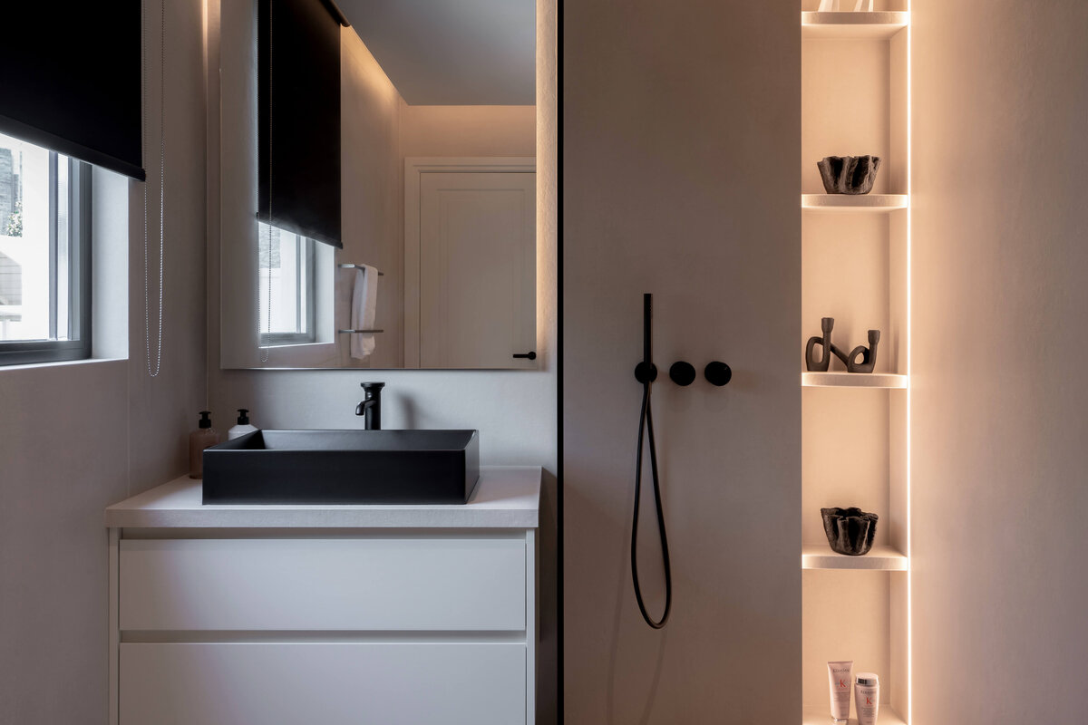 Modern, minimal bathroom with a sink on one side and a softly lit walk-in shower with black hardware on the right.