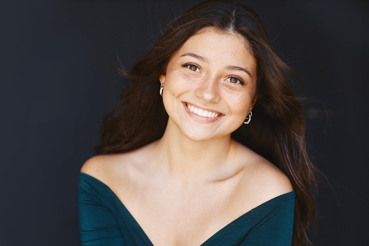 High school senior girl smiling with windblown hair in an off-the-shoulder emerald top against a black studio backdrop. Clean, natural light headshot with confident and approachable expression.