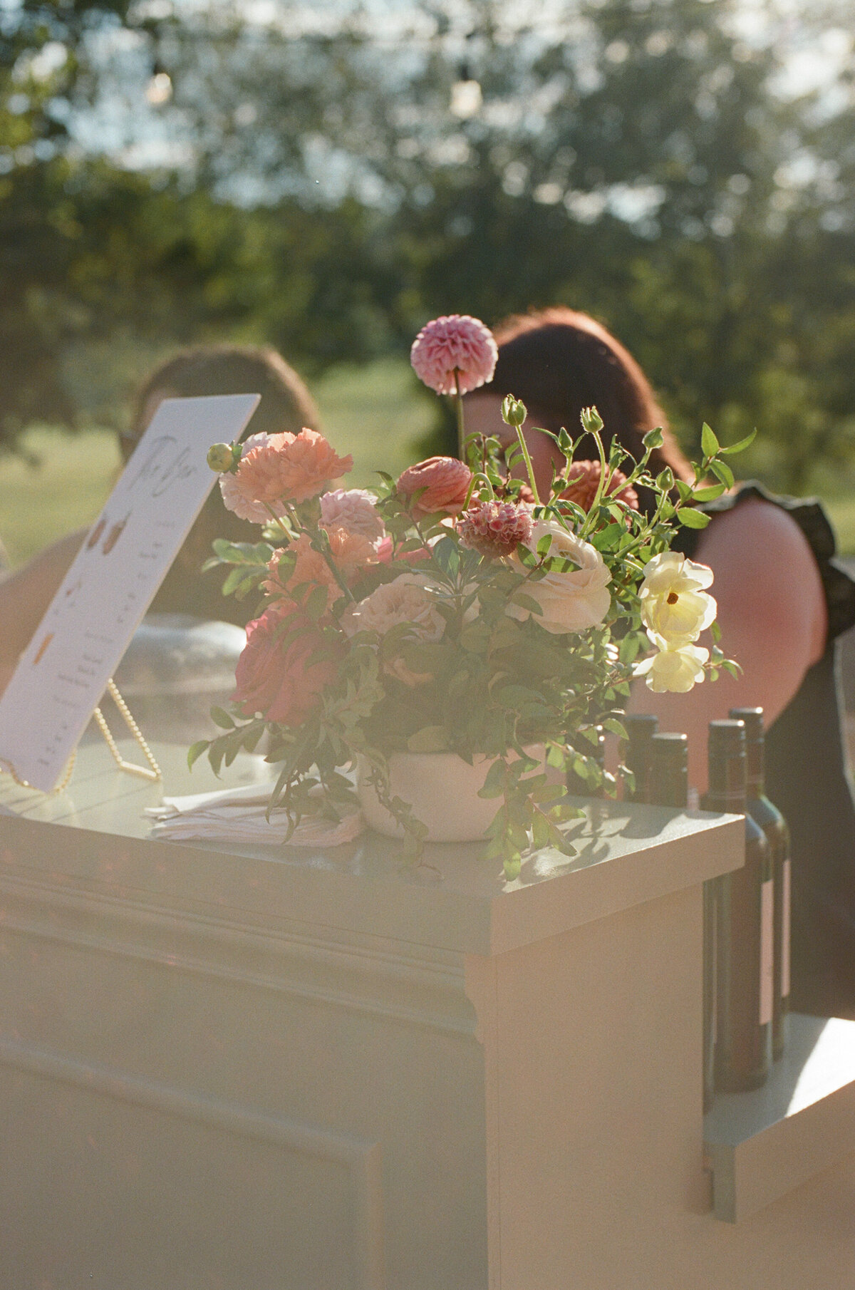 Vibrant floral centerpiece featuring dahlias, roses, and ranunculus displayed on an outdoor wedding bar during cocktail hour, styled with fresh greenery for a colorful garden-inspired celebration.
