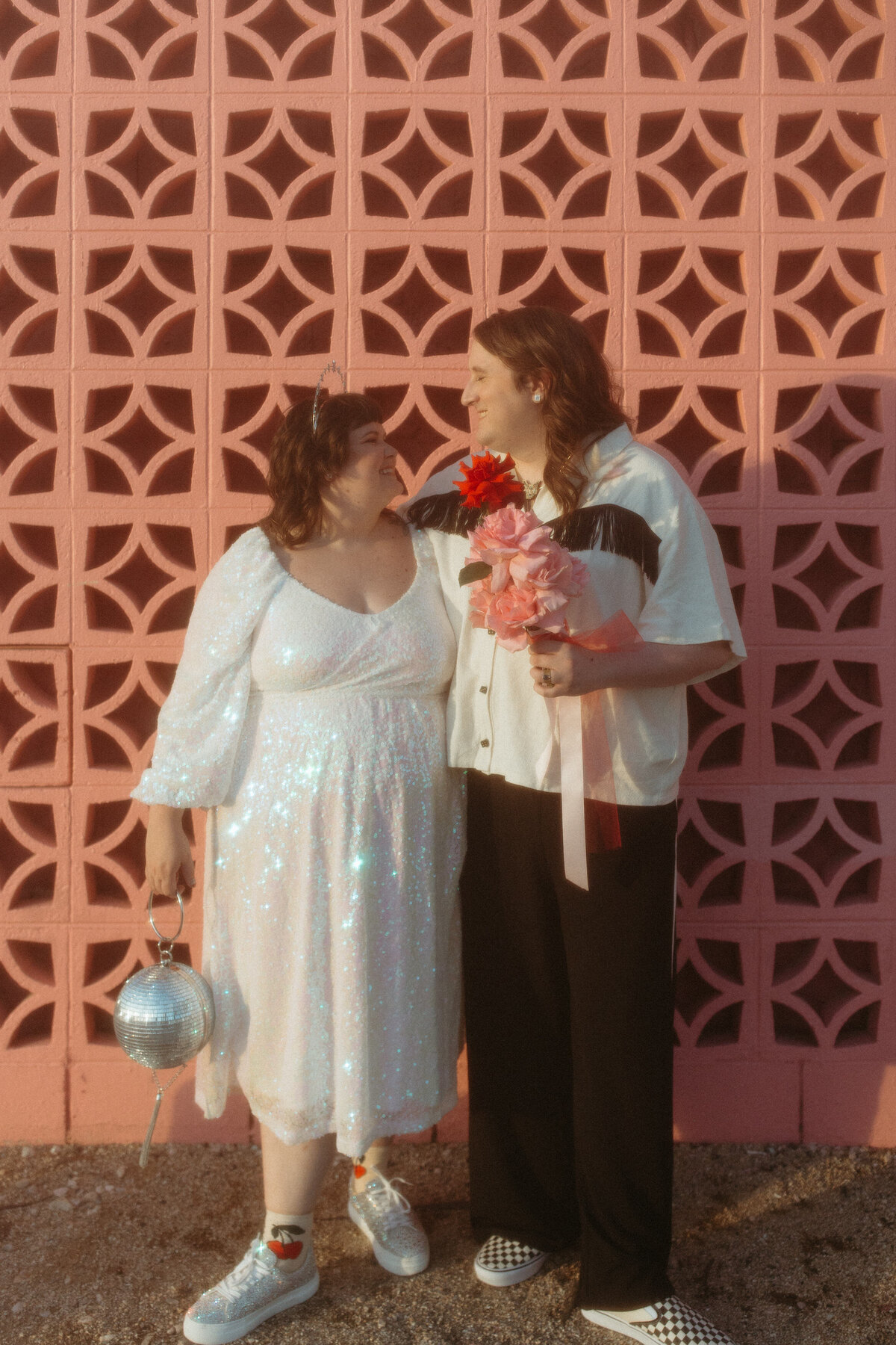 Two people in casual wedding attire exchanging a loving glance in front of a pink geometric wall, one holding a disco ball purse and a bouquet of red flowers