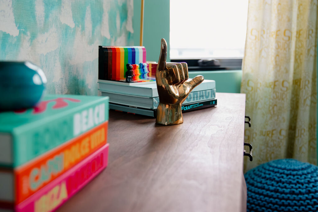 A close-up shot of the top surface of a mid century modern dresser. Several stacks of colorful books are arranged on the desk. On the left, a stack features a teal book on top with the word "BEACH" visible. In the center, a stack of light blue books is topped with a small, colorful striped object and a shiny gold-colored hand sculpture making the "hang loose" shaka sign. In the background, a window with patterned curtains allows soft light to enter. A portion of a teal knitted pouf is visible in the bottom right corner. The wall behind the desk has a textured light turquoise and white pattern.