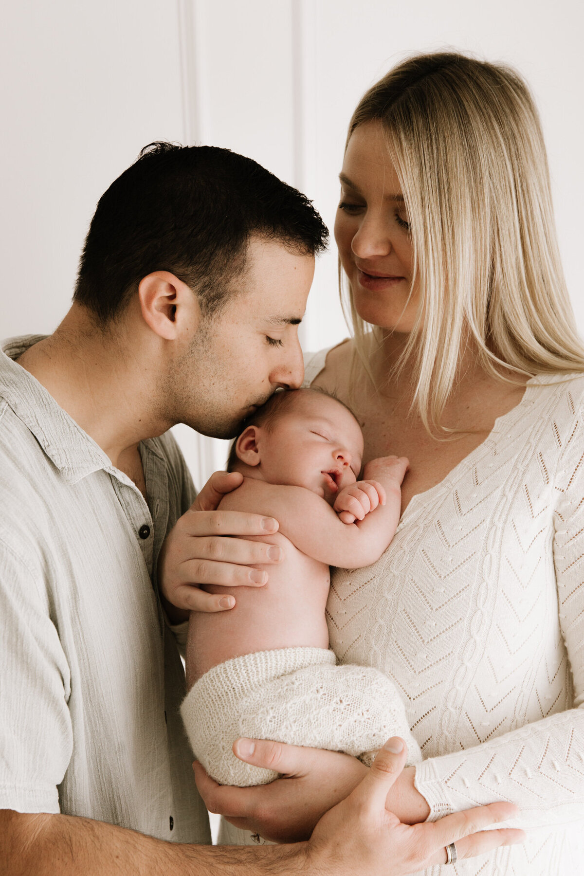 Close-up portrait of parents holding and cuddling their sleeping newborn baby in a softly lit Sydney photography studio, radiating warmth and love.