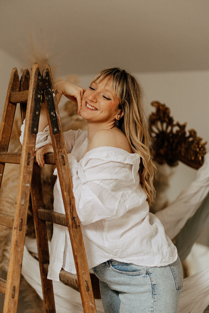 Femme blonde en chemise blanche et jean, coude posé sur un escabeau prise au studio en Vendée.