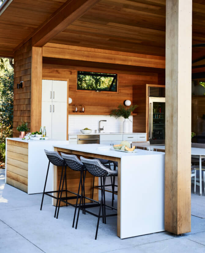 Kitchen designers in Boston create an outdoor kitchen with light grey cabinets, red cedar accents, and a high-top table with luxury European furniture in Hingham, MA.