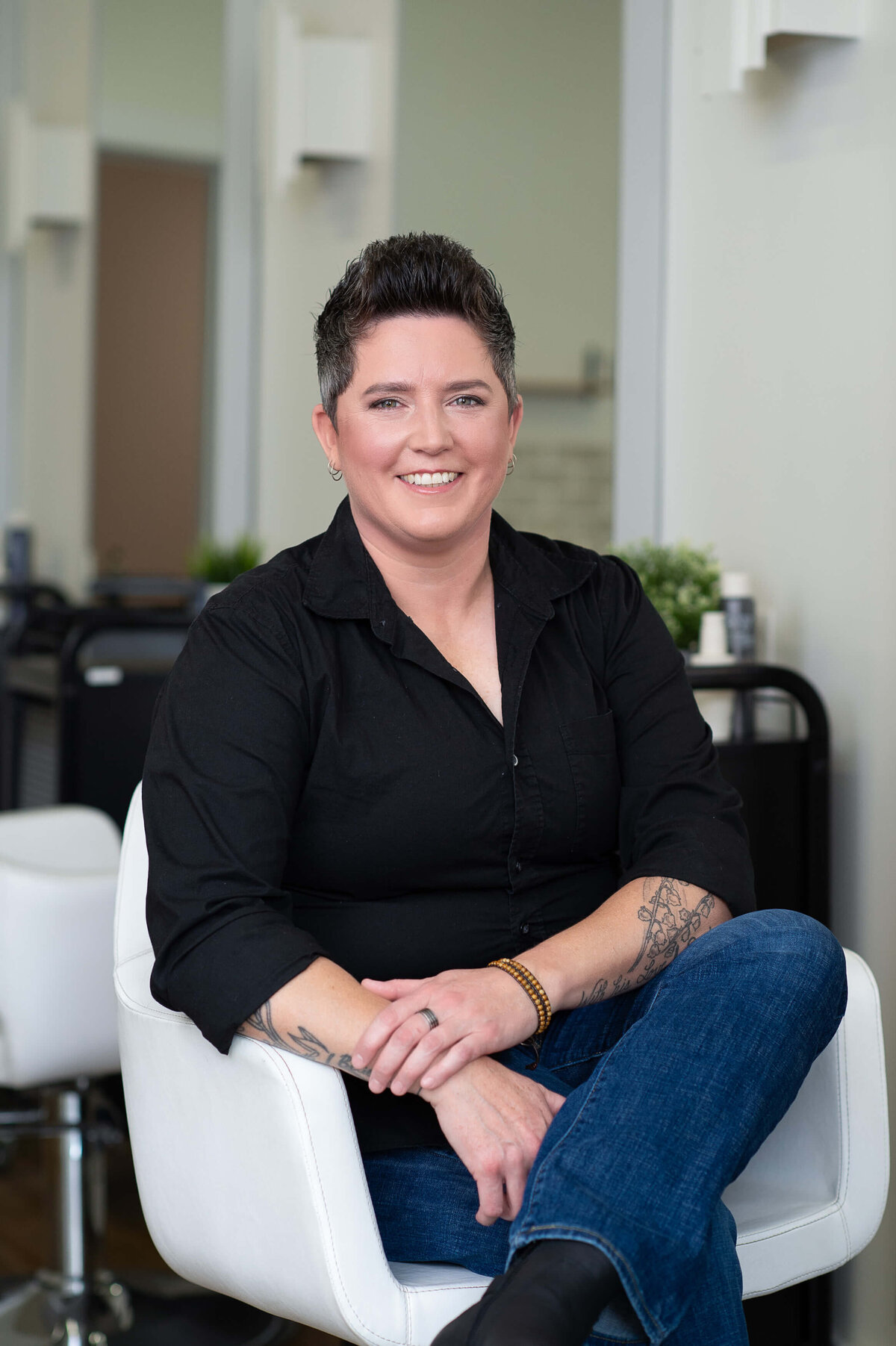 a woman with short dark hair, black top and jeans sitting in a white chair.  Captured on location at Richardson Hair Design by Ottawa branding photographer JEMMAN Photography