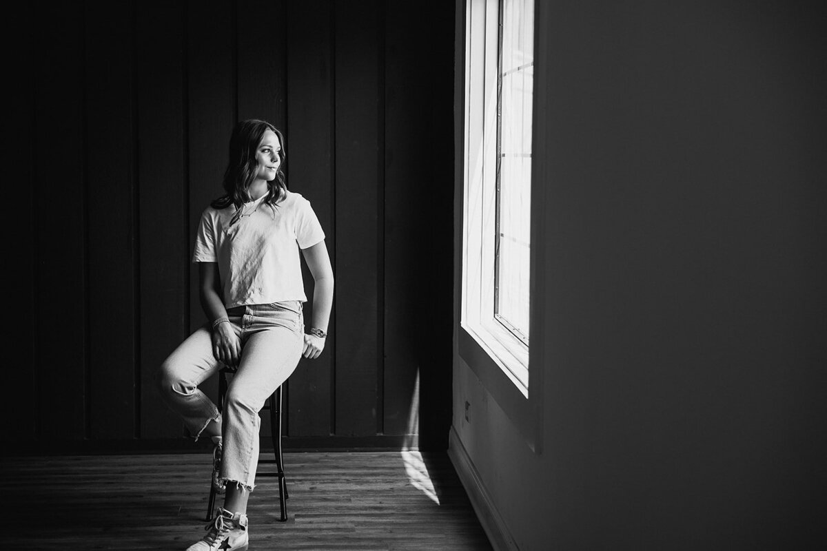Black and white senior portrait of a girl sitting on a stool looking out of a window