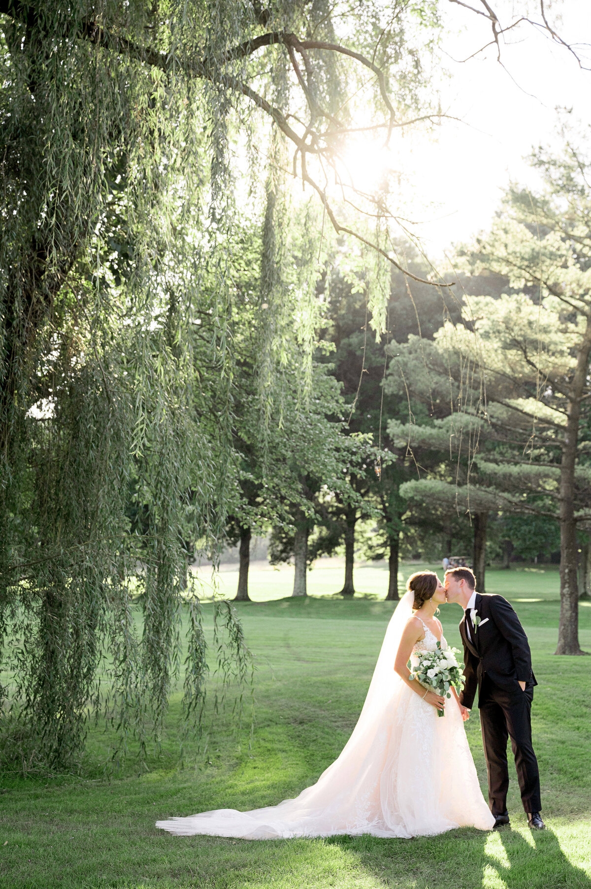 bride and groom kissing during their wedding at westmoreland golf club standing on the green grass with a weeping willow tree