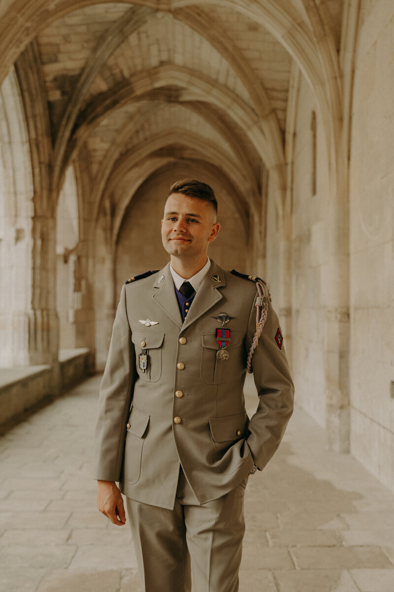 Marié en uniforme posant souriant et main dans la poche sous des voûtes en pierre pour Laura, lors d'une séance photo mariage.