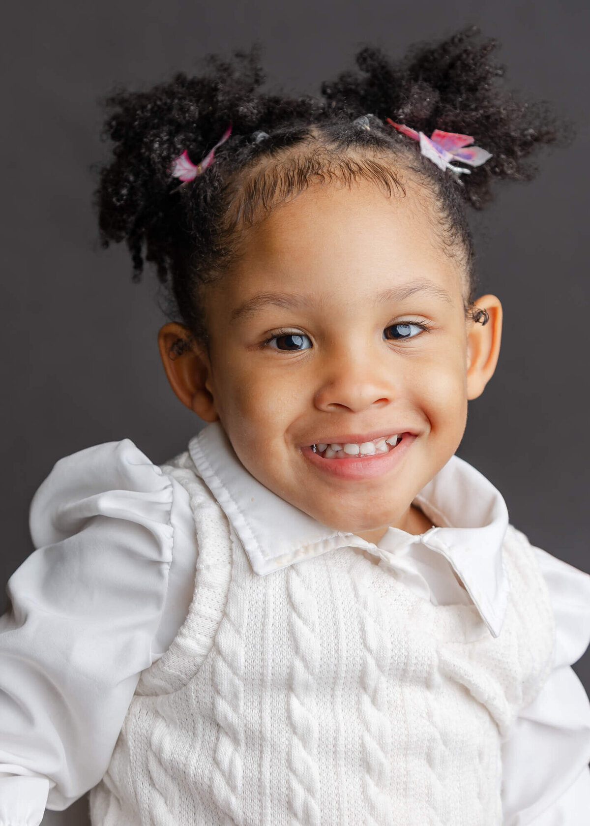 School portrait of a preschool age black girl with gurly pigtails in her hair smiling  and wearing a white shirt and sweater on a gray background