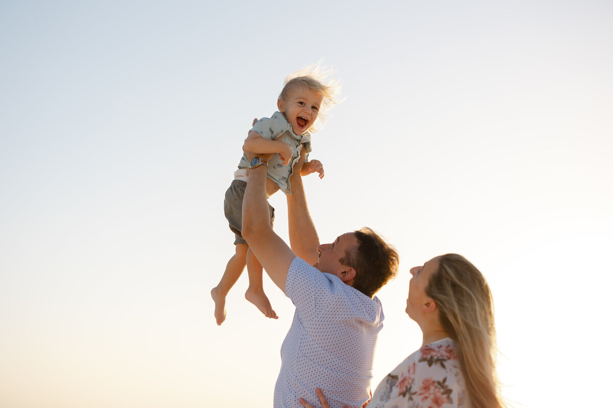 a father swings his laughing child into the sunset sky as the mother laughs behind him
