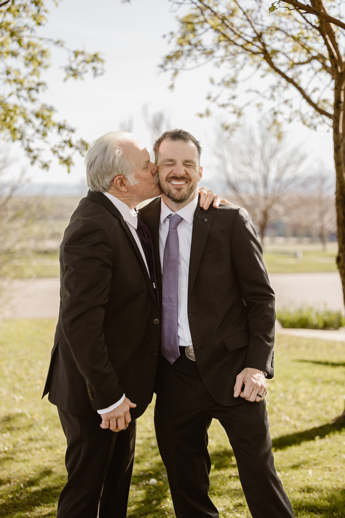 Older man in a suit affectionately kisses a smiling groom on the cheek during an outdoor wedding, with sunlight filtering through spring trees in the background.