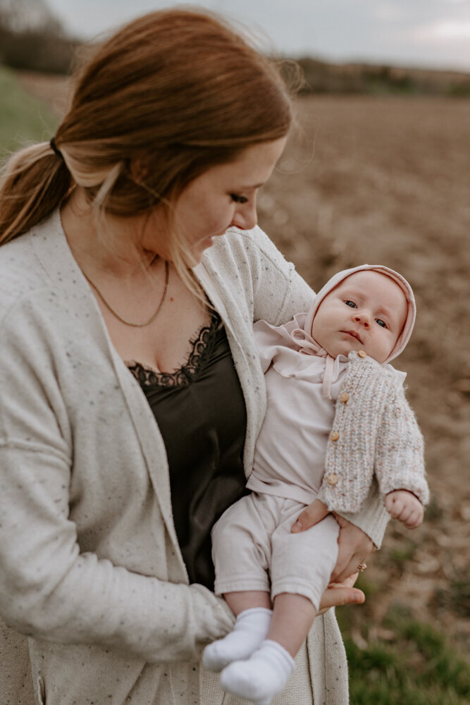 Close up image. Mom holding infant daughter in her arms for a family photoshoot in Exeter, Ontario. The baby is looking at the camera, the mom is looking at the baby and slightly blurred.